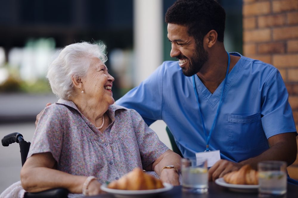An elderly woman in a wheelchair is sitting at a table with a nurse.