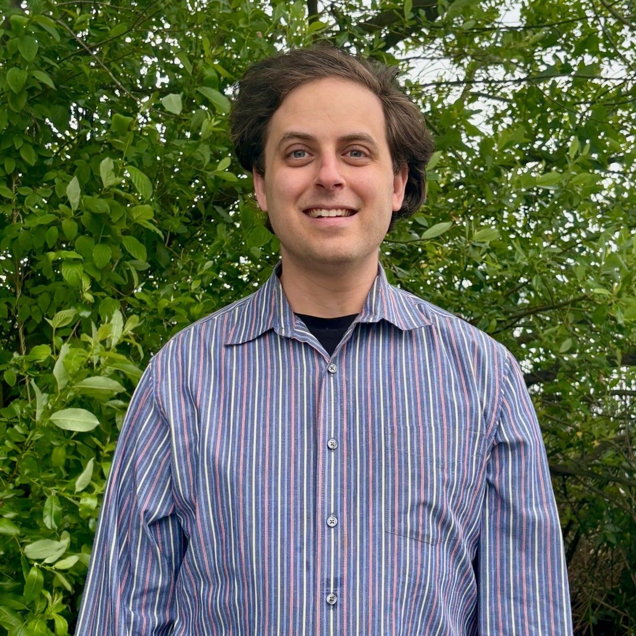 A man in a striped shirt is standing in front of trees. A man in a striped shirt is standing in front of trees.