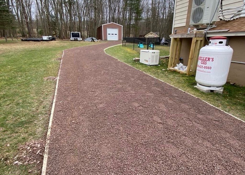 Red gravel driveway leading toward a garage and yard, next to a house with propane tank.