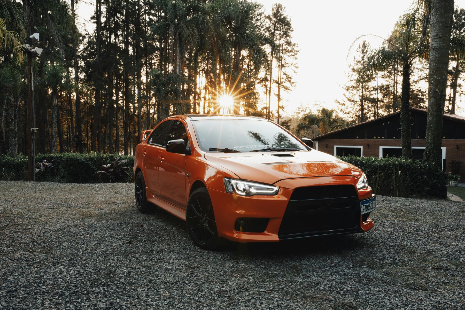 Orange sports car parked on gravel, with sun setting behind trees.