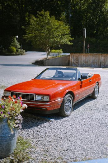 Red convertible parked on gravel, next to a flower pot, in a sunny outdoor setting.