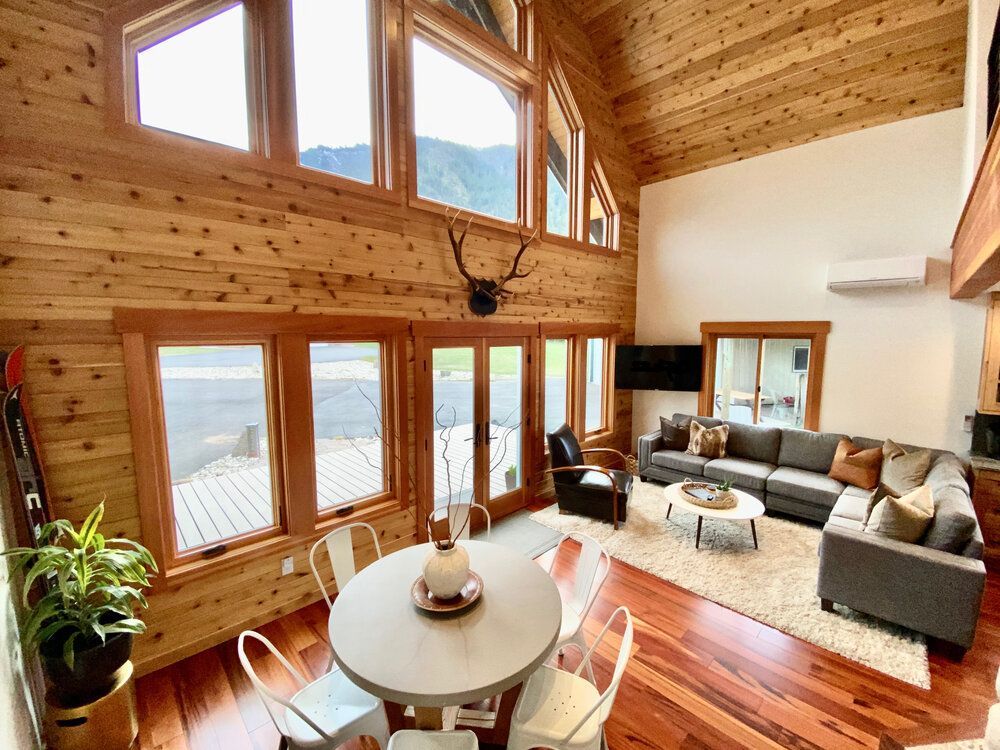 A overview of a living room with a couch, and a dining room and deer antlers on the wall.