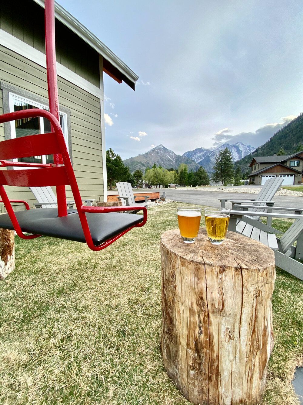 Two glasses of beer are sitting on a wooden stump next to a chair swing in front of a house.