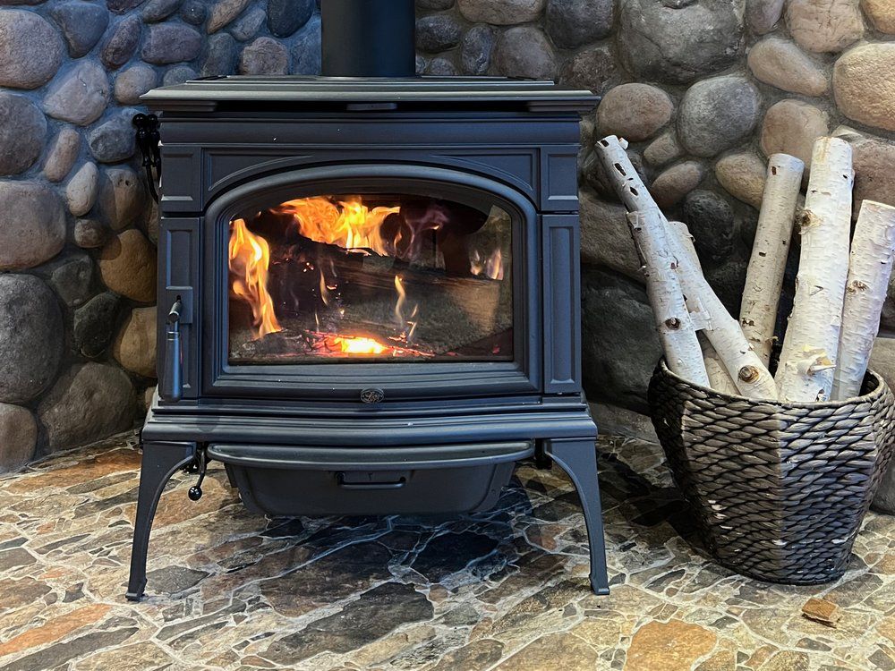 A lit wood stove is sitting in front of a stone wall next to a basket of logs.