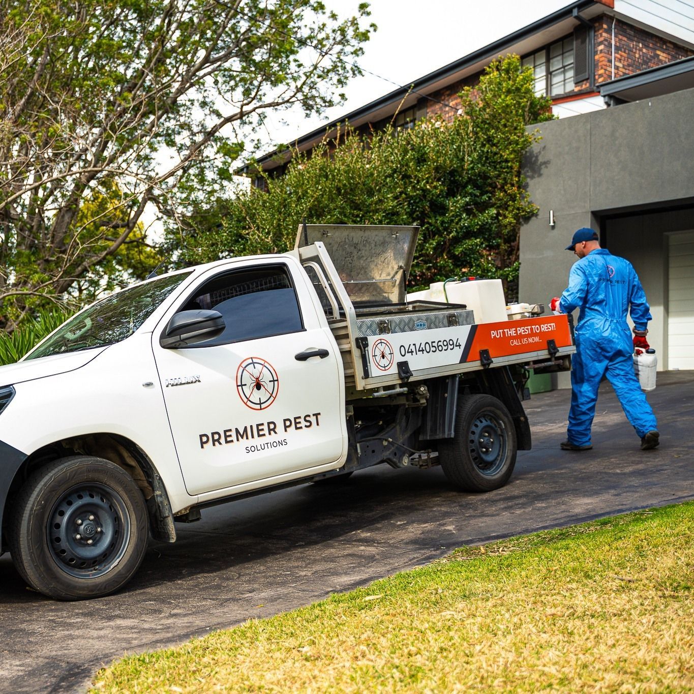 White pest control truck in driveway; worker in blue uniform walks toward house with a bucket — Premier Pest Control Wollongong In Dapto, NSW