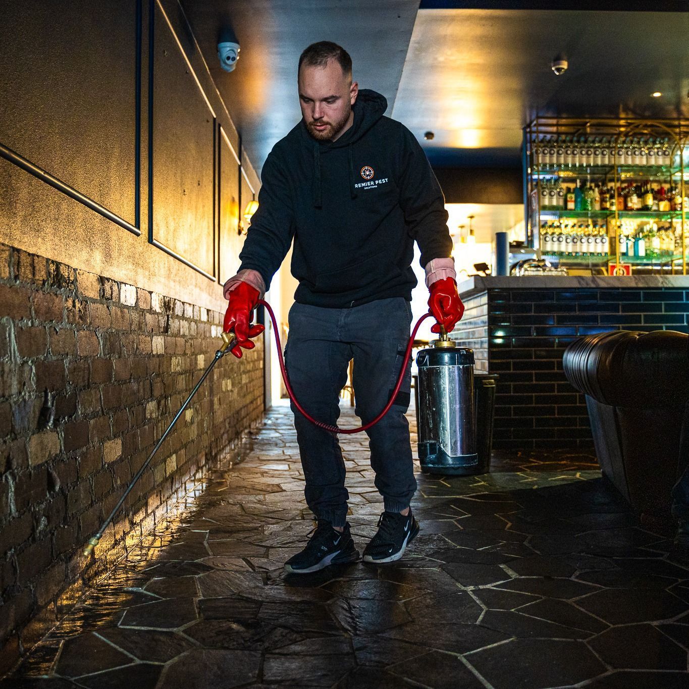 Man in a hooded sweatshirt spraying a brick wall and floor with a red hose in a bar — Premier Pest Control Wollongong In Kiama, NSW