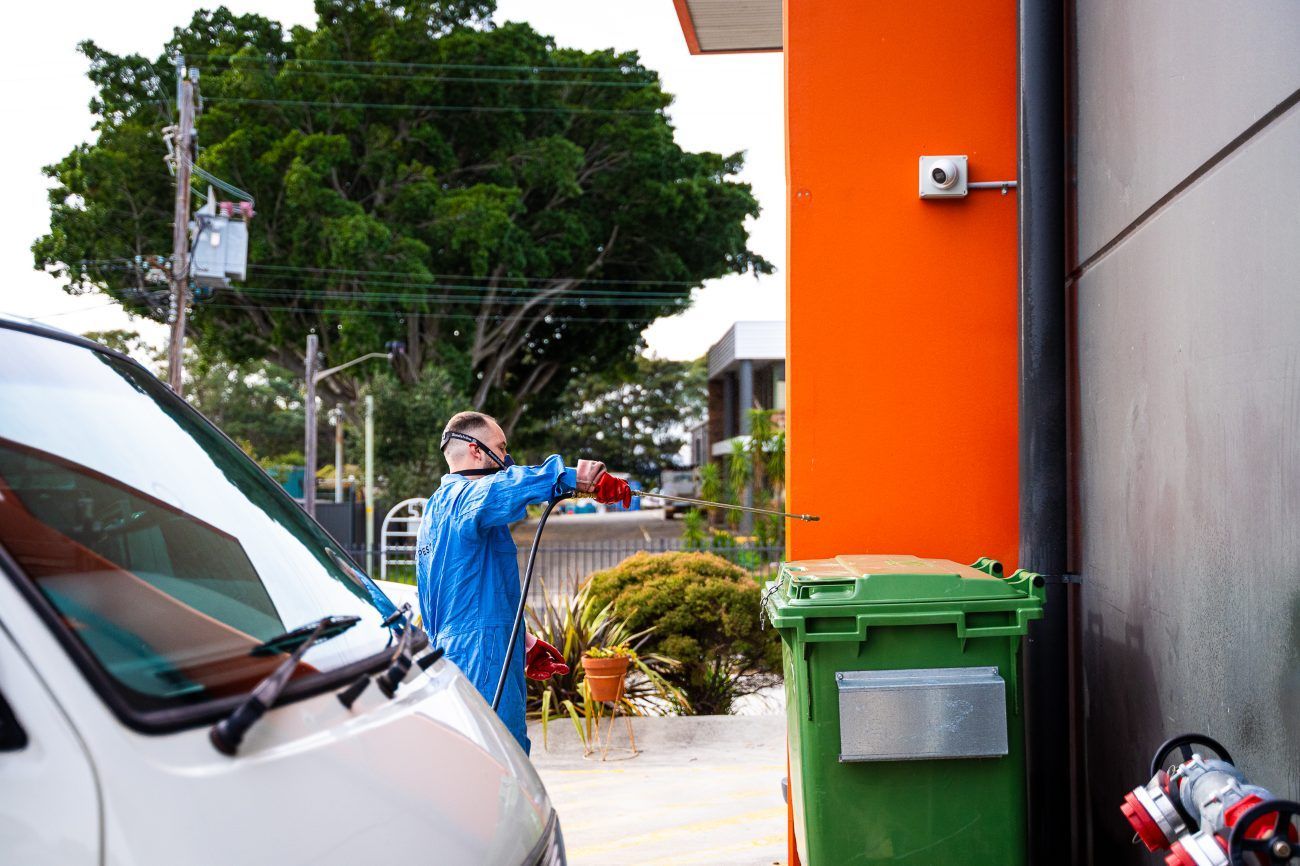 man in blue pest control suit is spraying a house next to a green bin and white van — Premier Pest Control Wollongong In Wollongong, NSW
