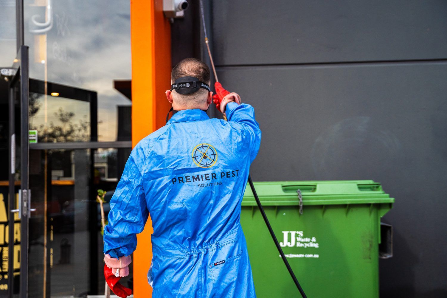 Man in blue jumpsuit spraying a building exterior near a green trash bin — Premier Pest Control Wollongong In Wollongong, NSW