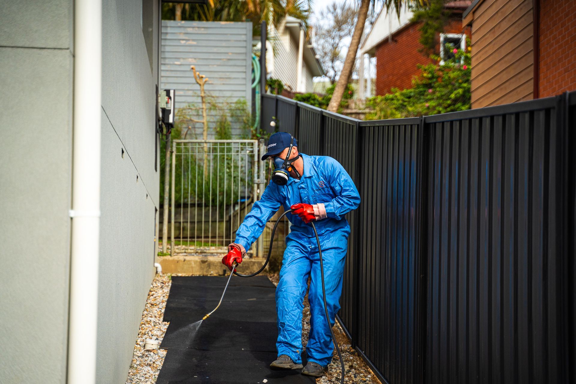 Pest control worker spraying pesticide along a wall and fence wearing a blue jumpsuit and respirator — Premier Pest Control Wollongong In Kiama, NSW
