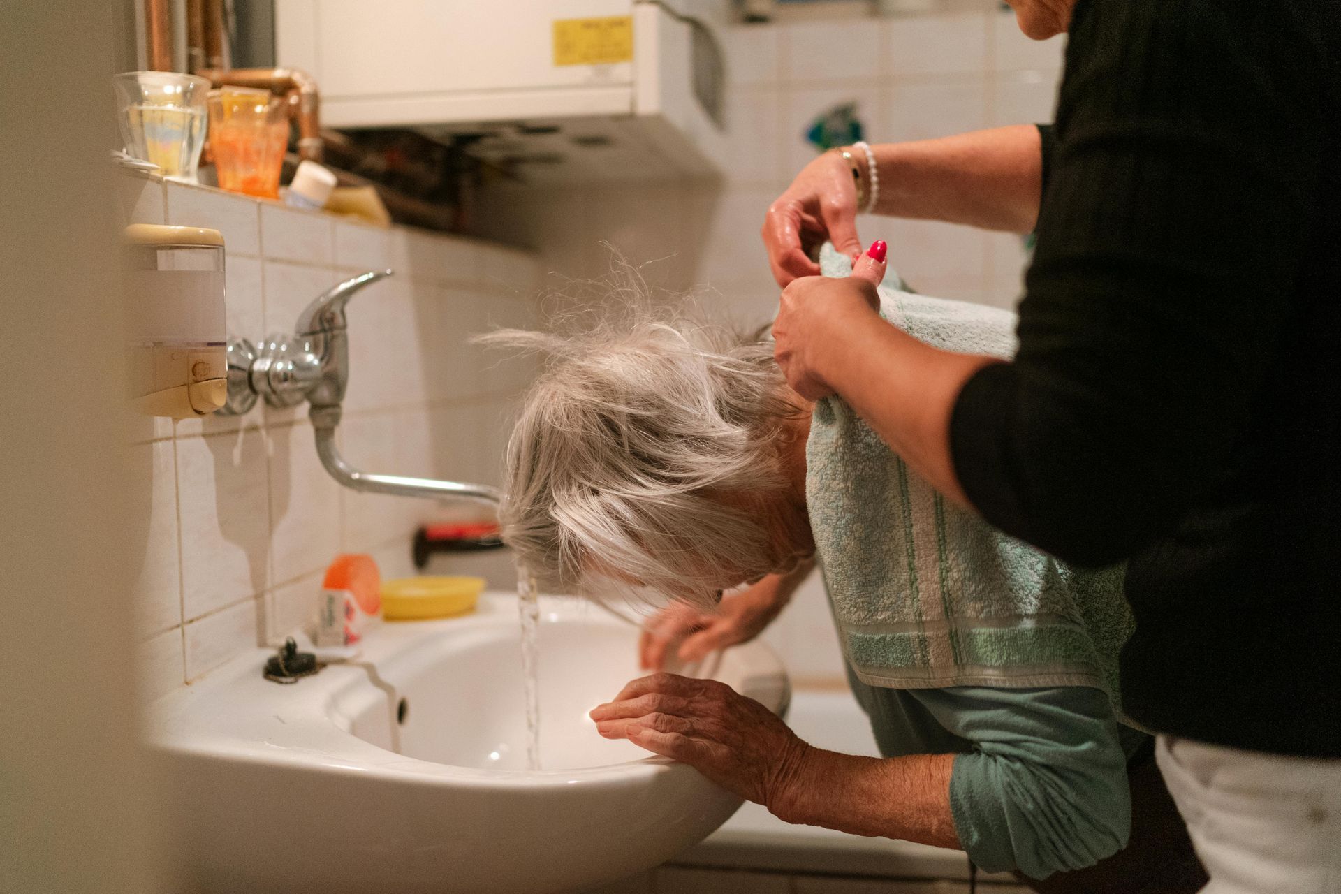 A woman is washing another woman 's hair in a bathroom sink.