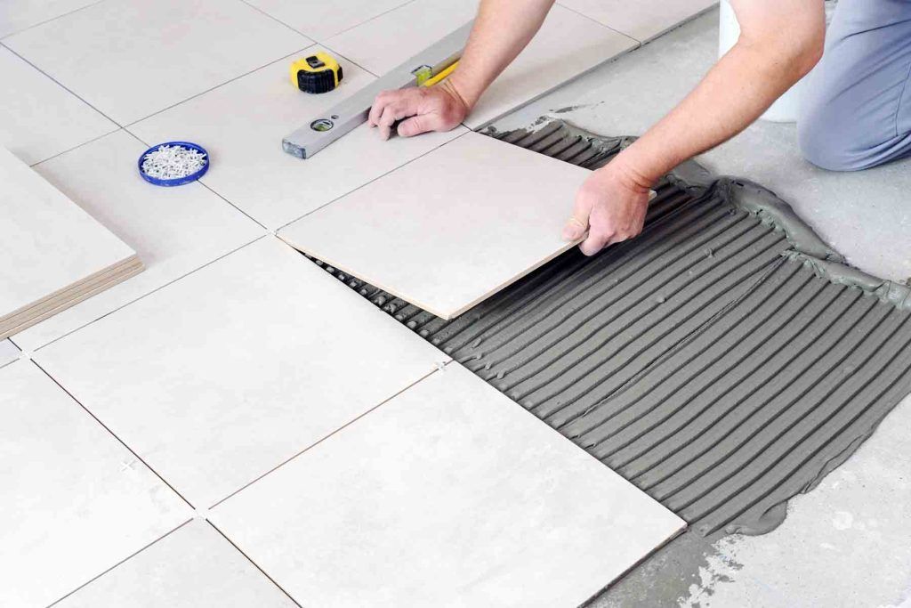 Person tiling a floor, applying a tile to a bed of gray mortar. Tools and tiles visible.