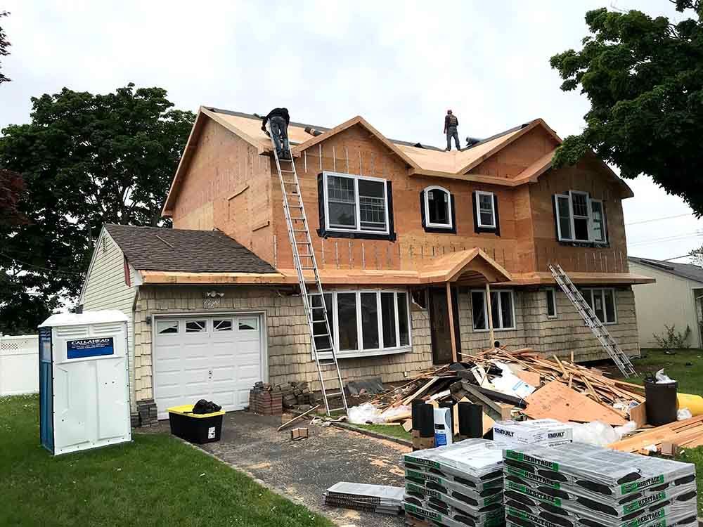 House under construction; roofers on ladders, wood framing visible, porta-potty on lawn.