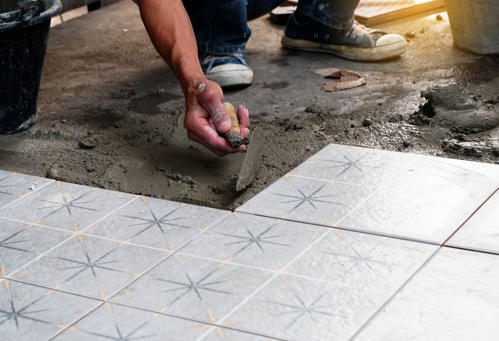Person laying tile, using a trowel to spread mortar on a floor, with a bucket and tiles in the background.