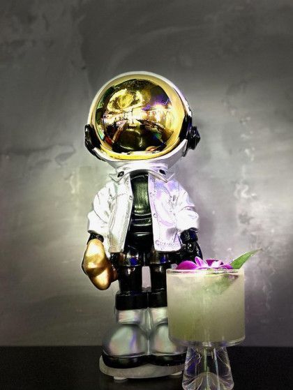 A statue of an astronaut is standing next to a drink on a table.