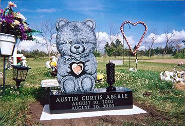 A child's grave marker. A large gray teddy bear sculpture holds a heart on a black granite base.