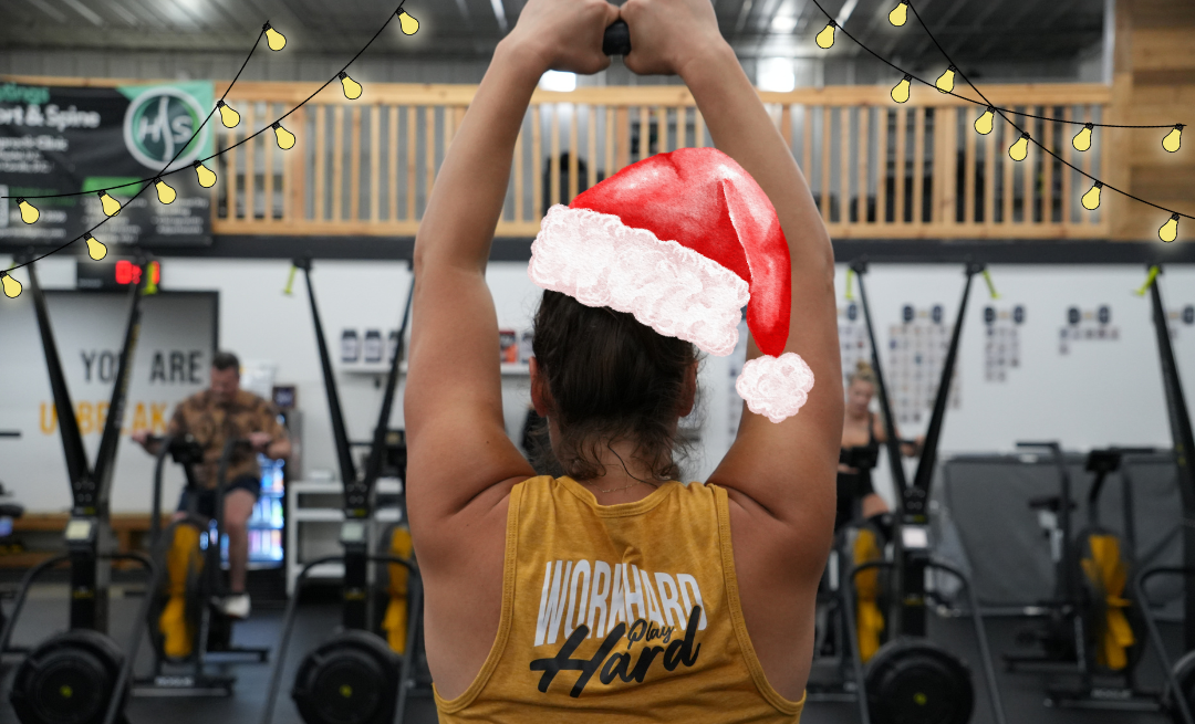 Person with Santa hat lifts kettlebell in gym, with other people working out. String lights overhead.