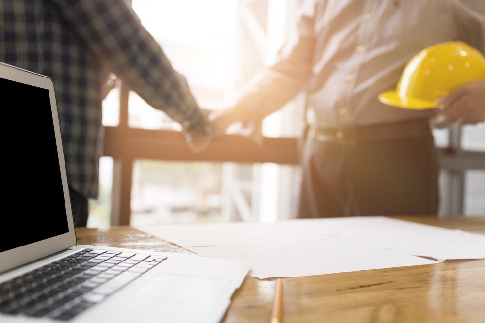 Two Men Shaking Hands Over A Table With A Laptop And A Hard Hat — Tradie Books Australia In Port Kennedy, WA