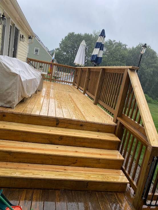 Wooden deck with stairs leading up, covered patio furniture, and two closed umbrellas on a rainy, overcast day.