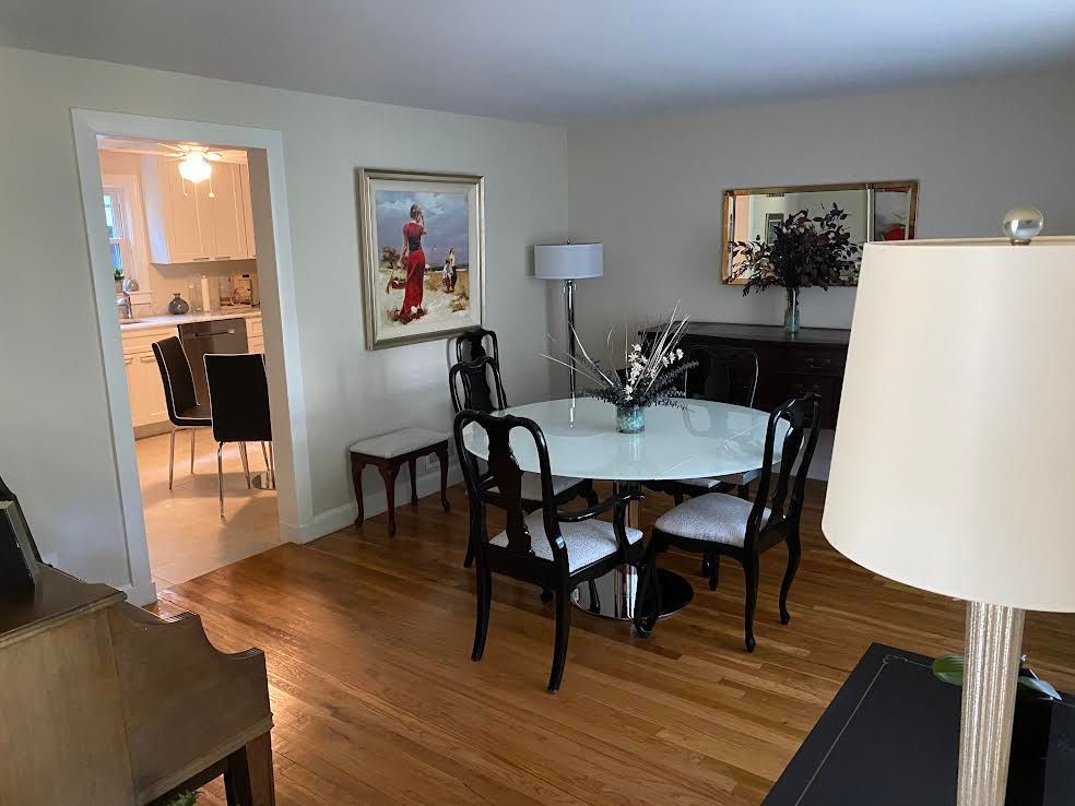 A dining room with a glass-topped table and four black chairs, hardwood floors, framed art, and a view into a kitchen.