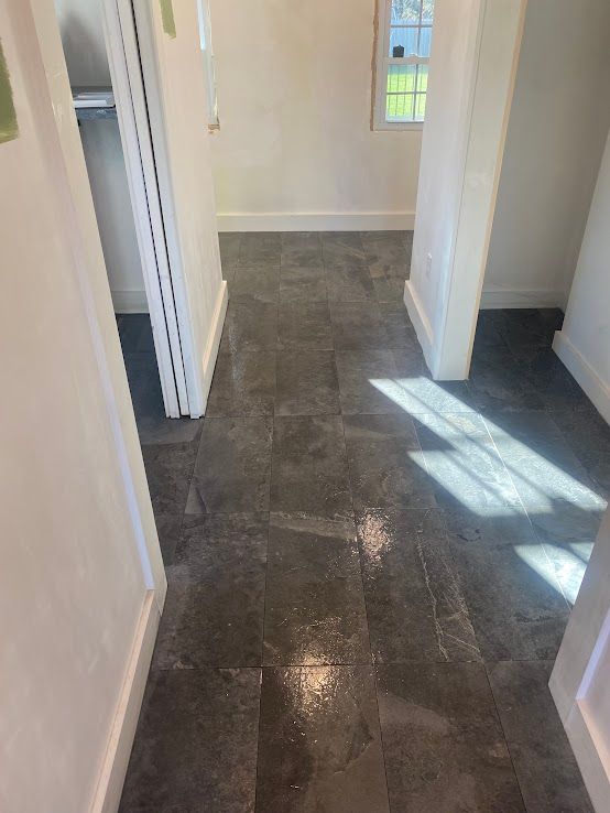 A hallway featuring dark gray stone tile flooring, white walls, and a window at the end, bathed in natural sunlight.