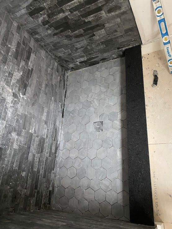 A shower stall under construction with black stone walls, gray hexagonal floor tiles, a center drain, and a level tool.