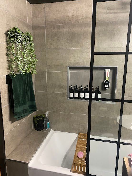 A modern bathroom with a bathtub, a recessed wall niche holding toiletries, a dark green towel, and hanging faux plants.