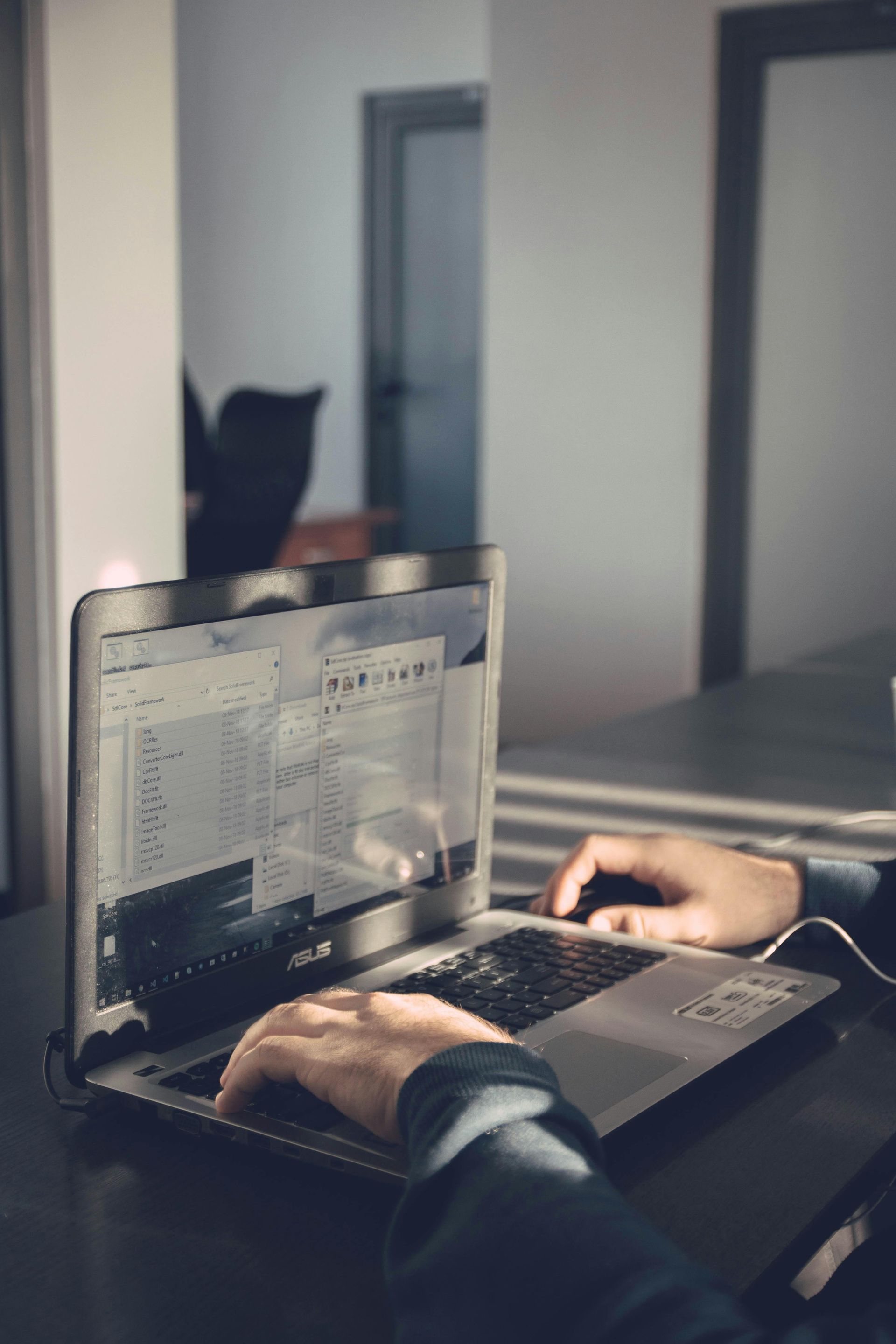 Hands typing on an open silver laptop on a desk in a dimly lit office.