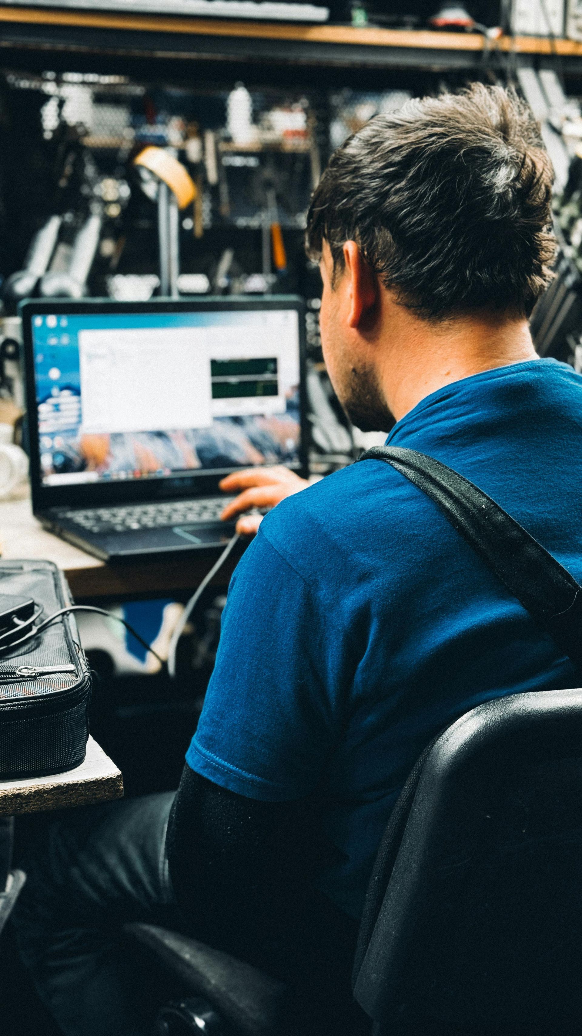 A person in a blue shirt works at a laptop in a cluttered, workshop-style setting.