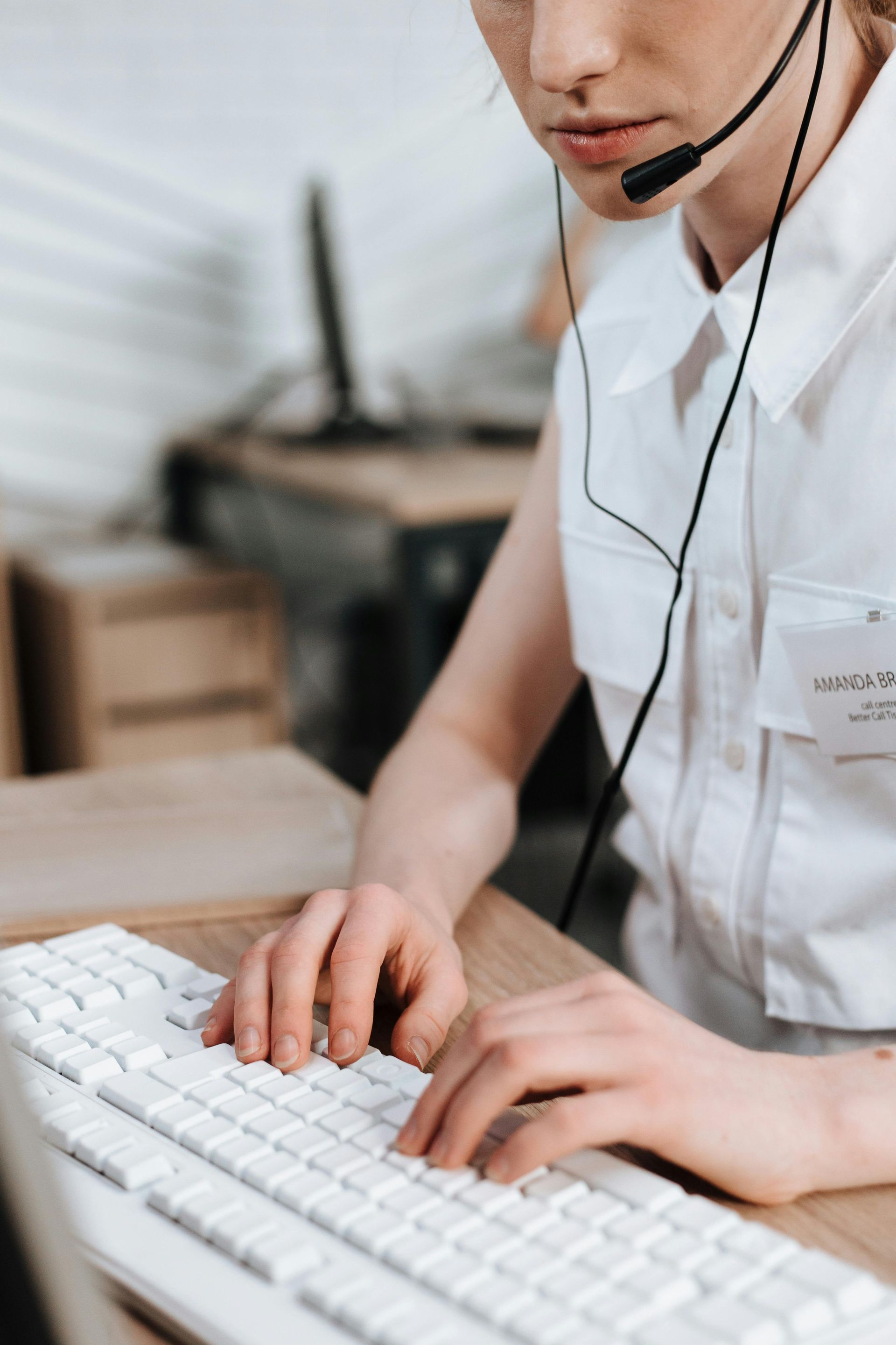 A person wearing a headset types on a white keyboard at a desk in an office setting.