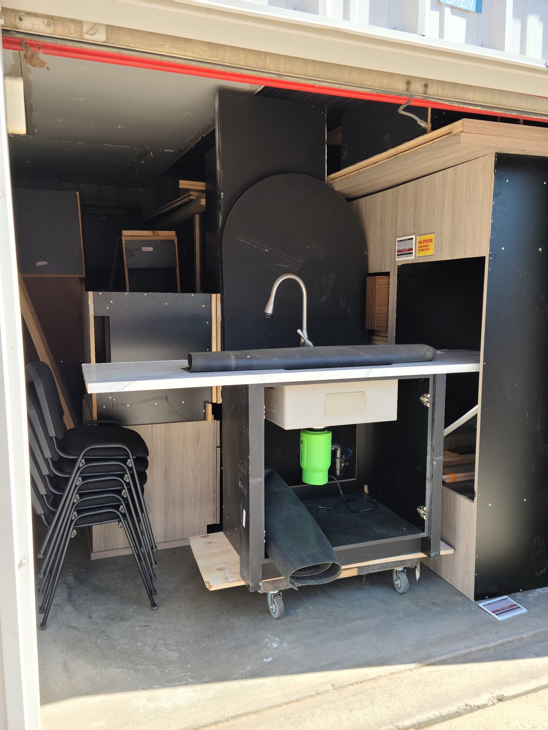 Inside a container, a mobile sink/counter unit with black and wood-tone elements, plus stacked chairs.