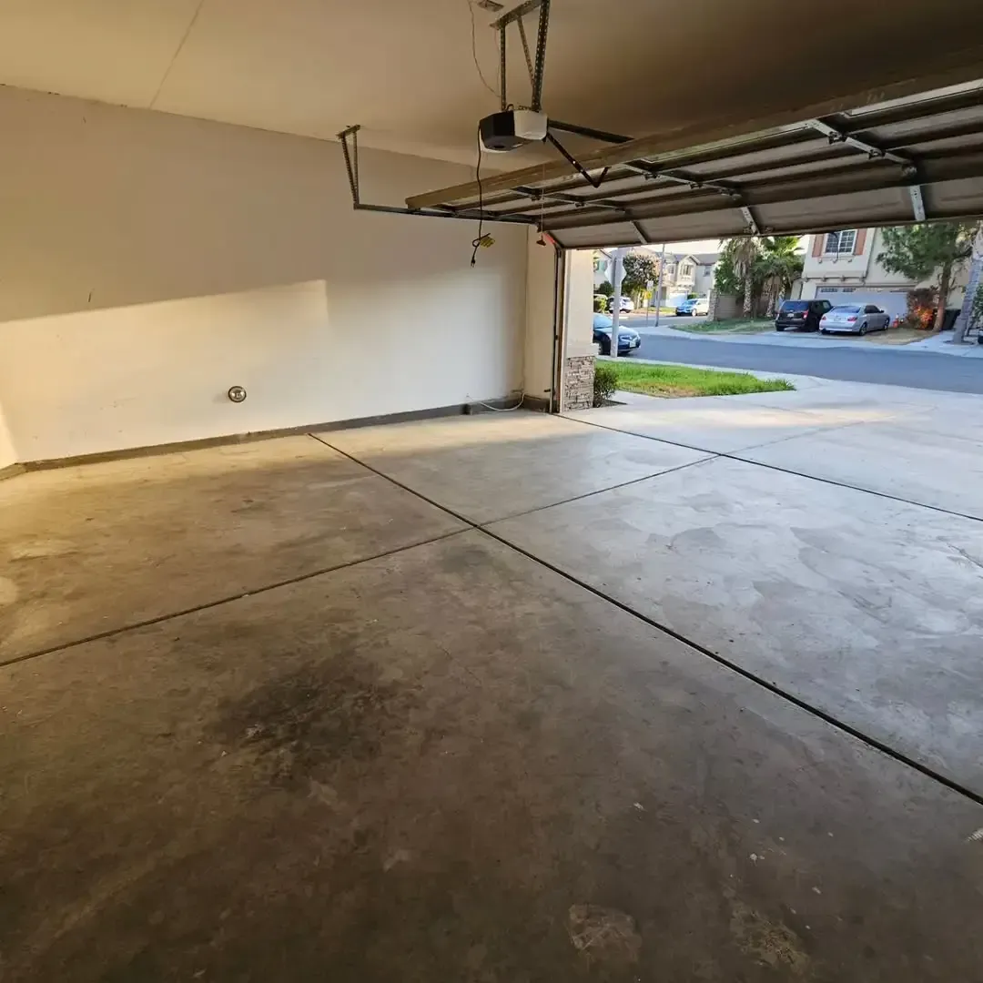 Empty garage with open door, view of street. Concrete floor, white walls, garage door opener.