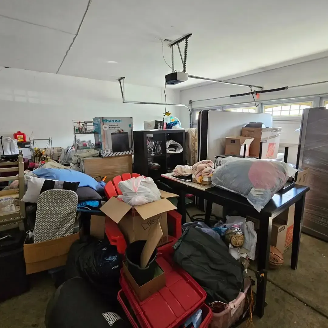 Cluttered garage with boxes, furniture, and various items piled up. The garage door is open.