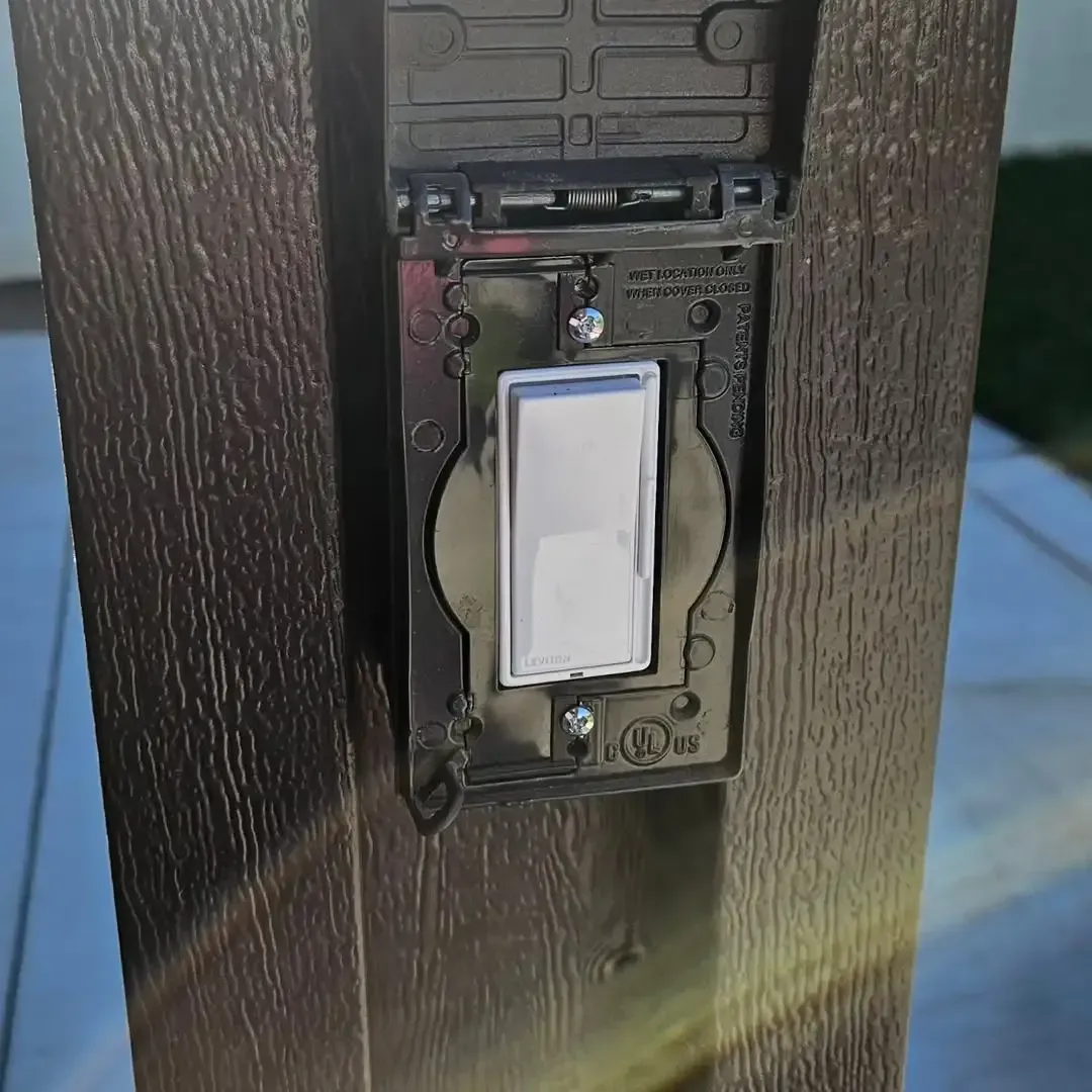 A black weatherproof electrical switch cover on a brown post; the switch is white and is on.