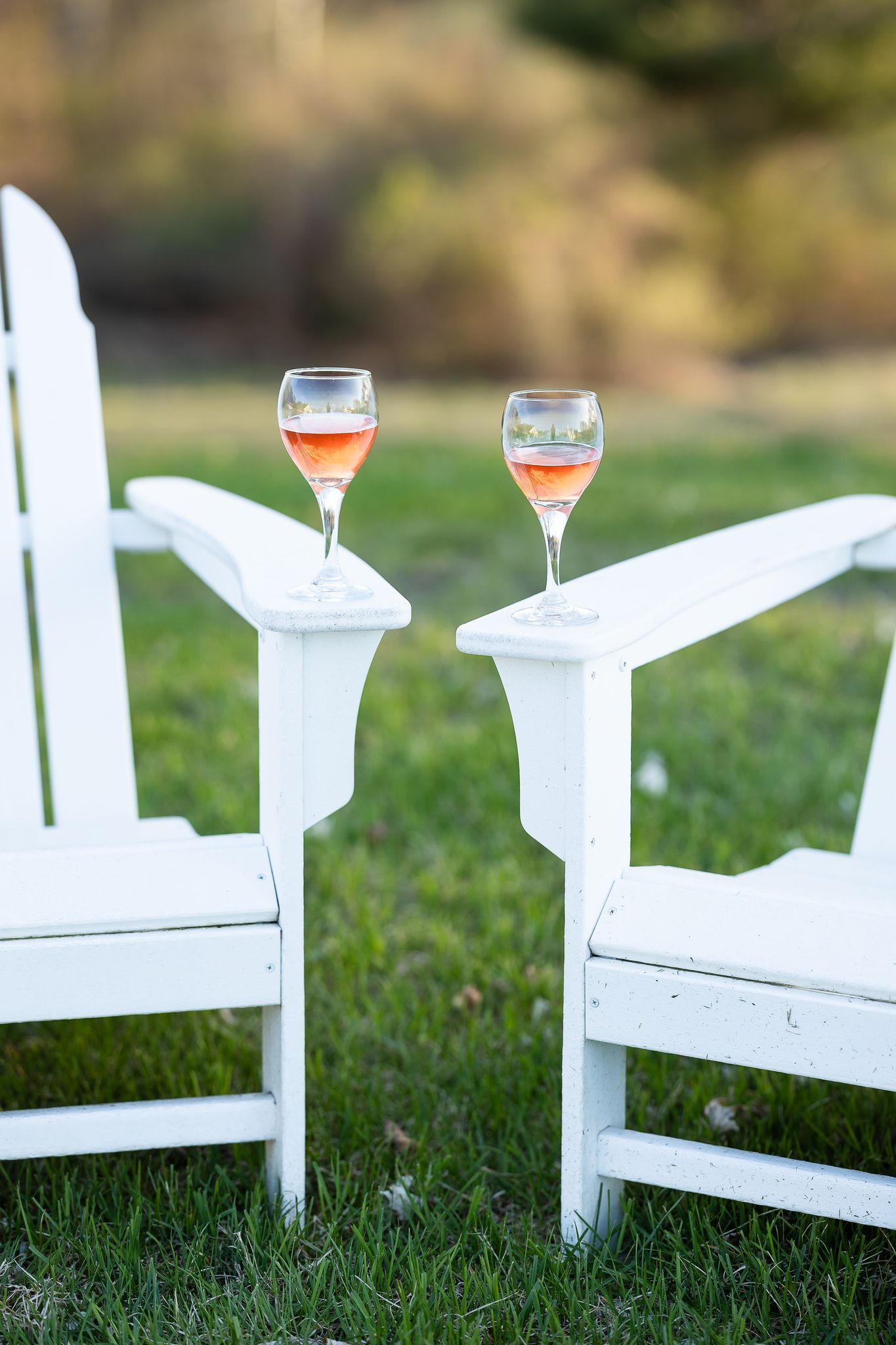 Two white Adirondack chairs with wine glasses on the armrests in the grass.