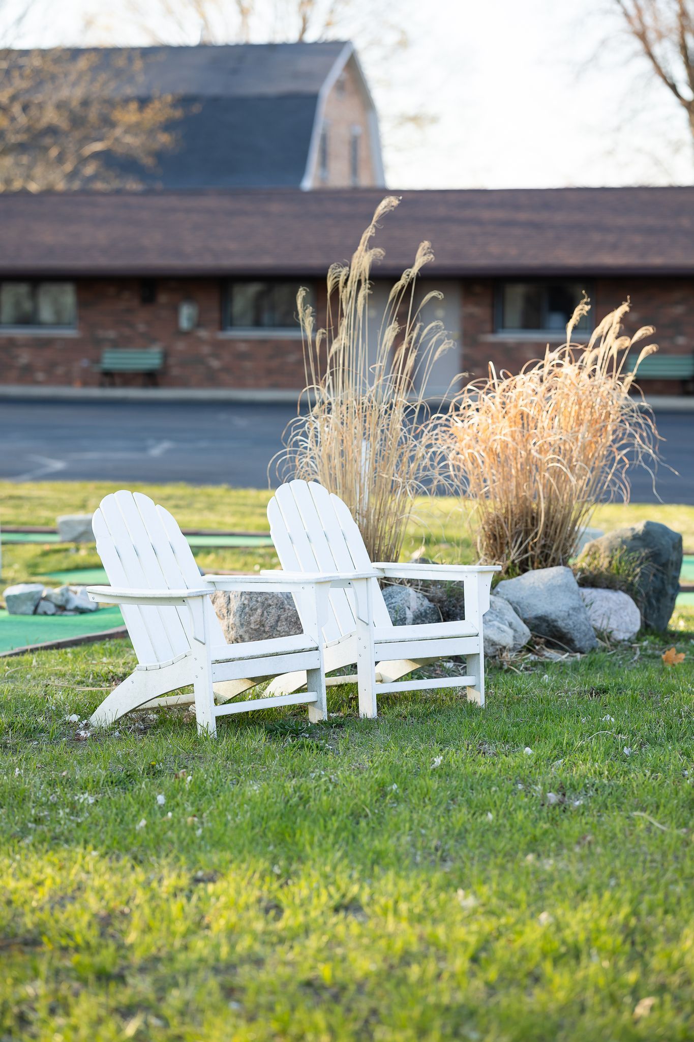 Two white Adirondack chairs sit in the grass in front of a house.