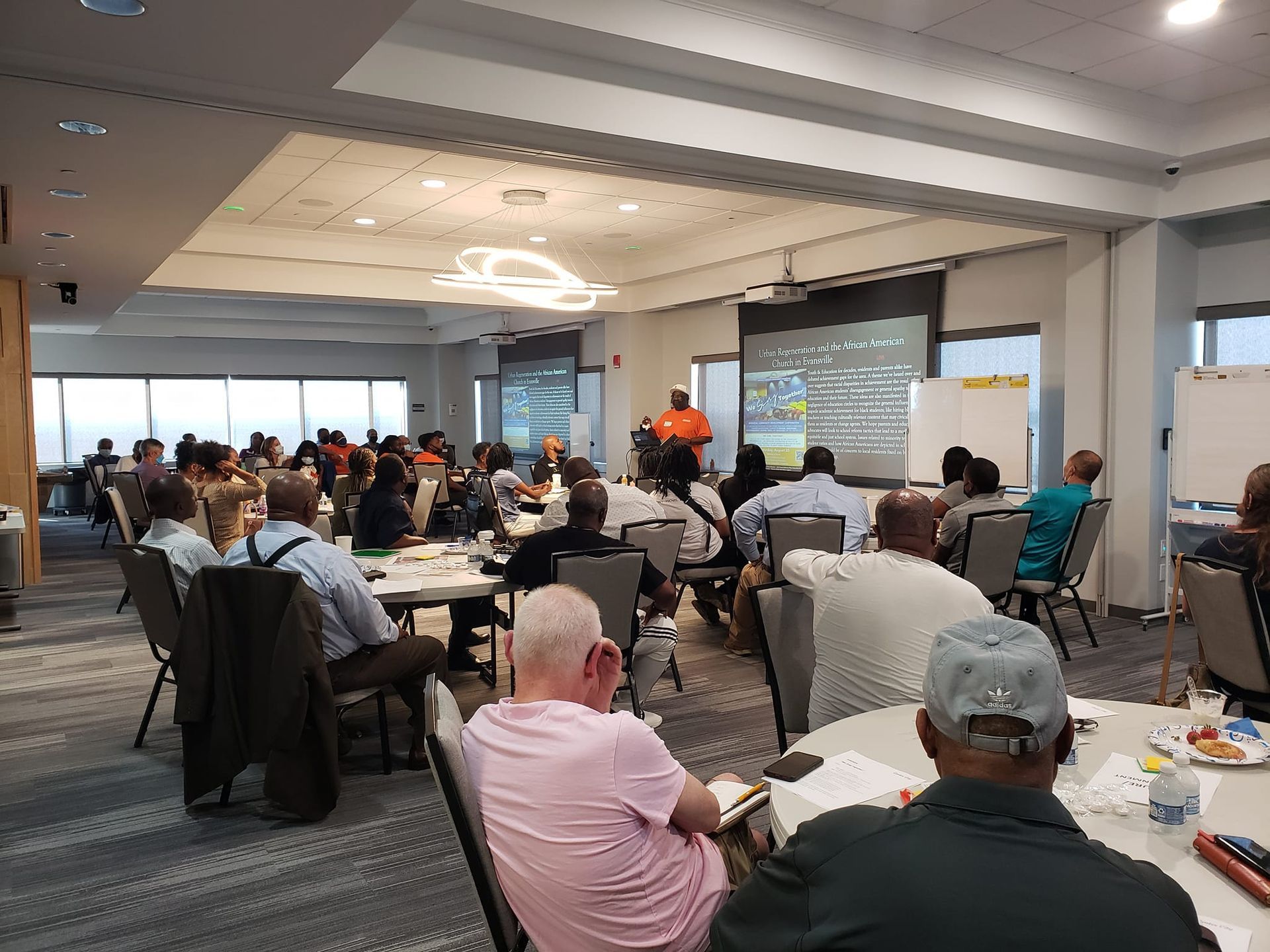 A group of people listens to a presentation in a modern conference room with a large screen and floor-to-ceiling windows.