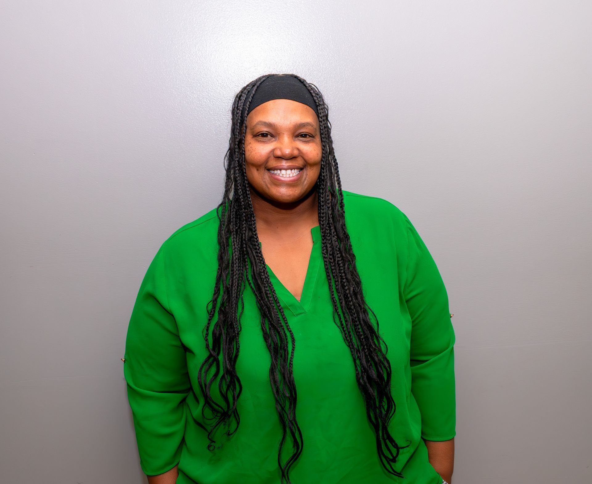 A person with long, dark braided hair smiles, wearing a green v-neck top against a plain light grey background.