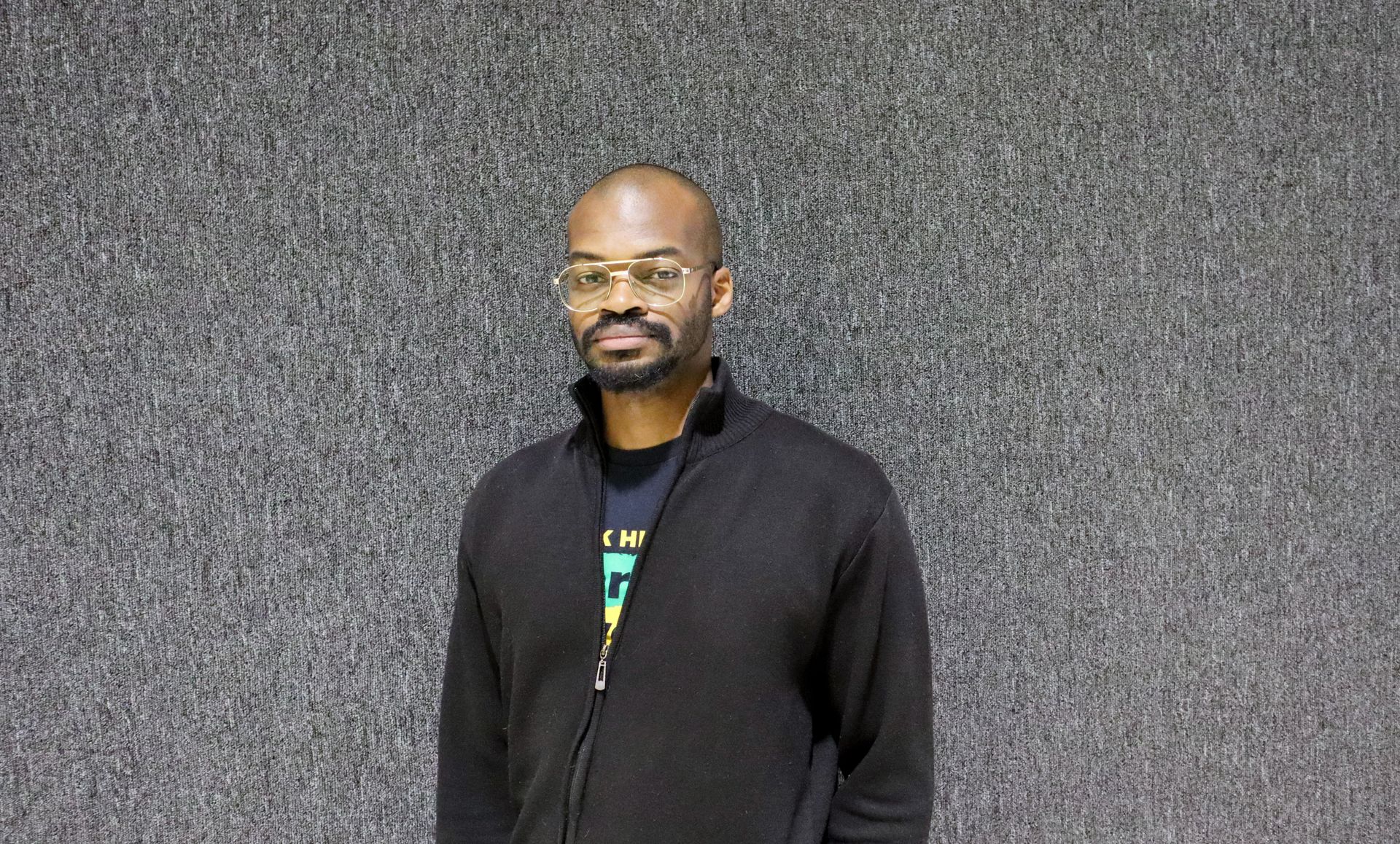 A person with short hair and glasses wearing a black zip-up sweater, positioned against a textured grey wall.