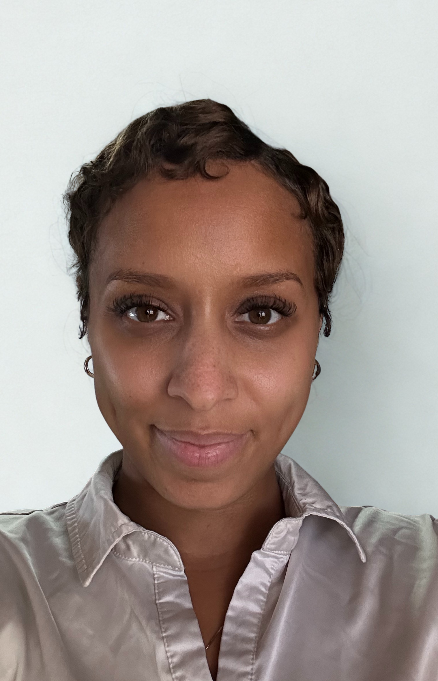 A person with a short, textured haircut wearing a silver collared shirt, posing against a light blue background.