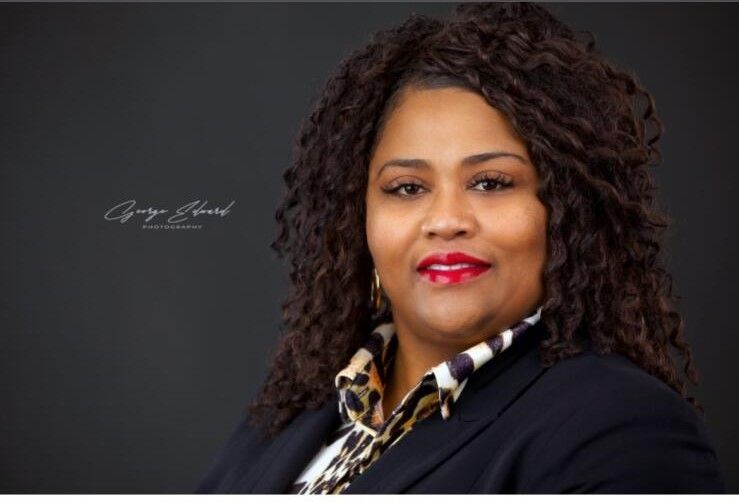 Headshot of a person with curly hair wearing a dark blazer over a leopard-print shirt against a dark background.