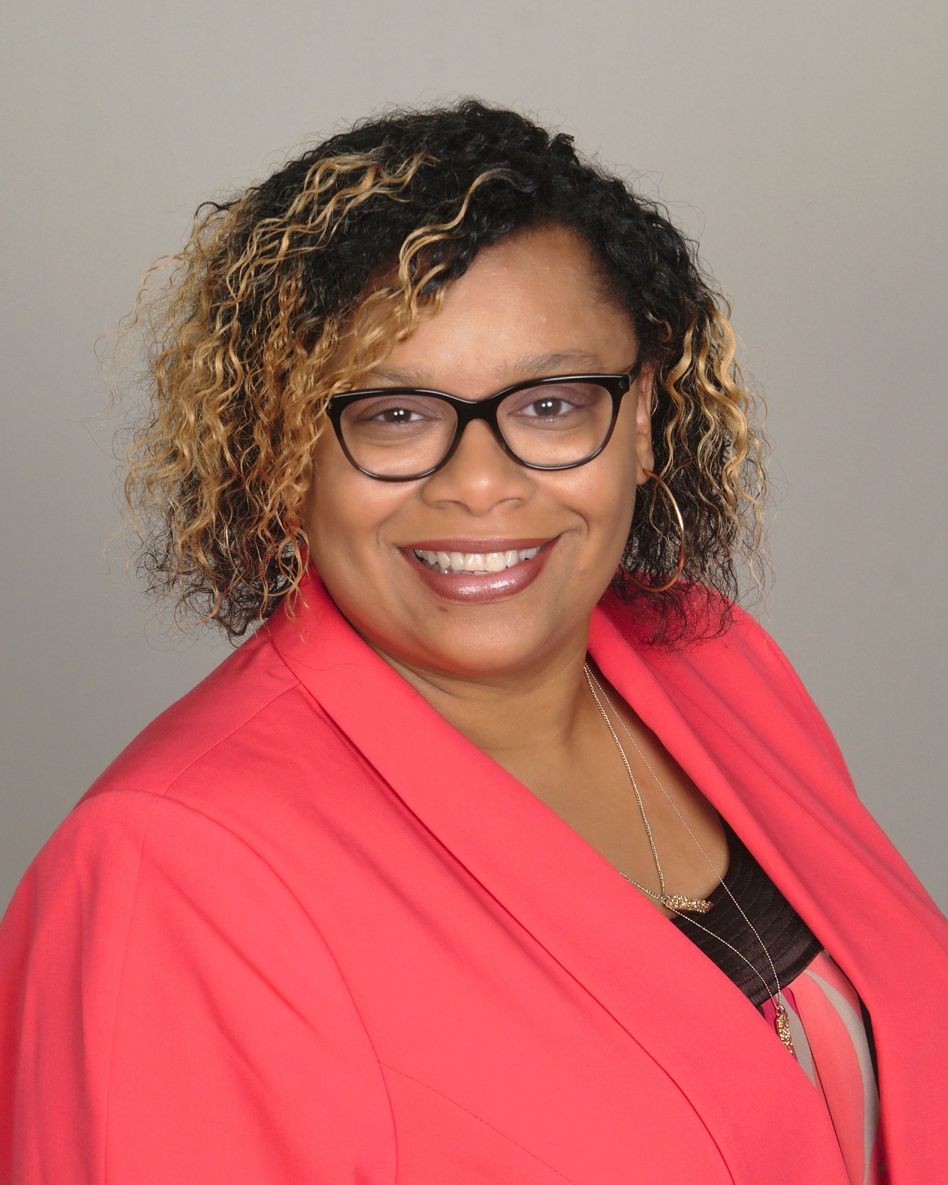 Smiling person wearing glasses and a bright pink blazer against a plain gray background.