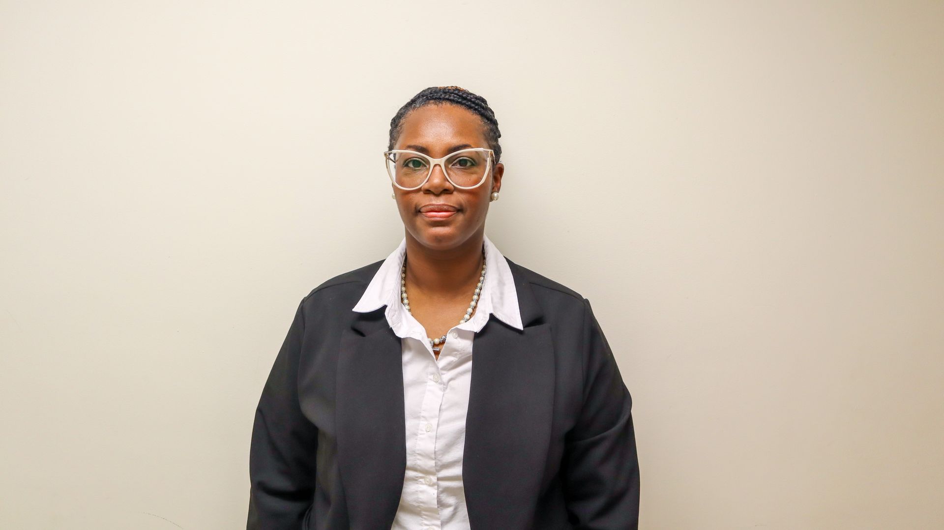 A person with short dark hair wearing glasses, a white collared shirt, and a dark blazer against a plain light background.
