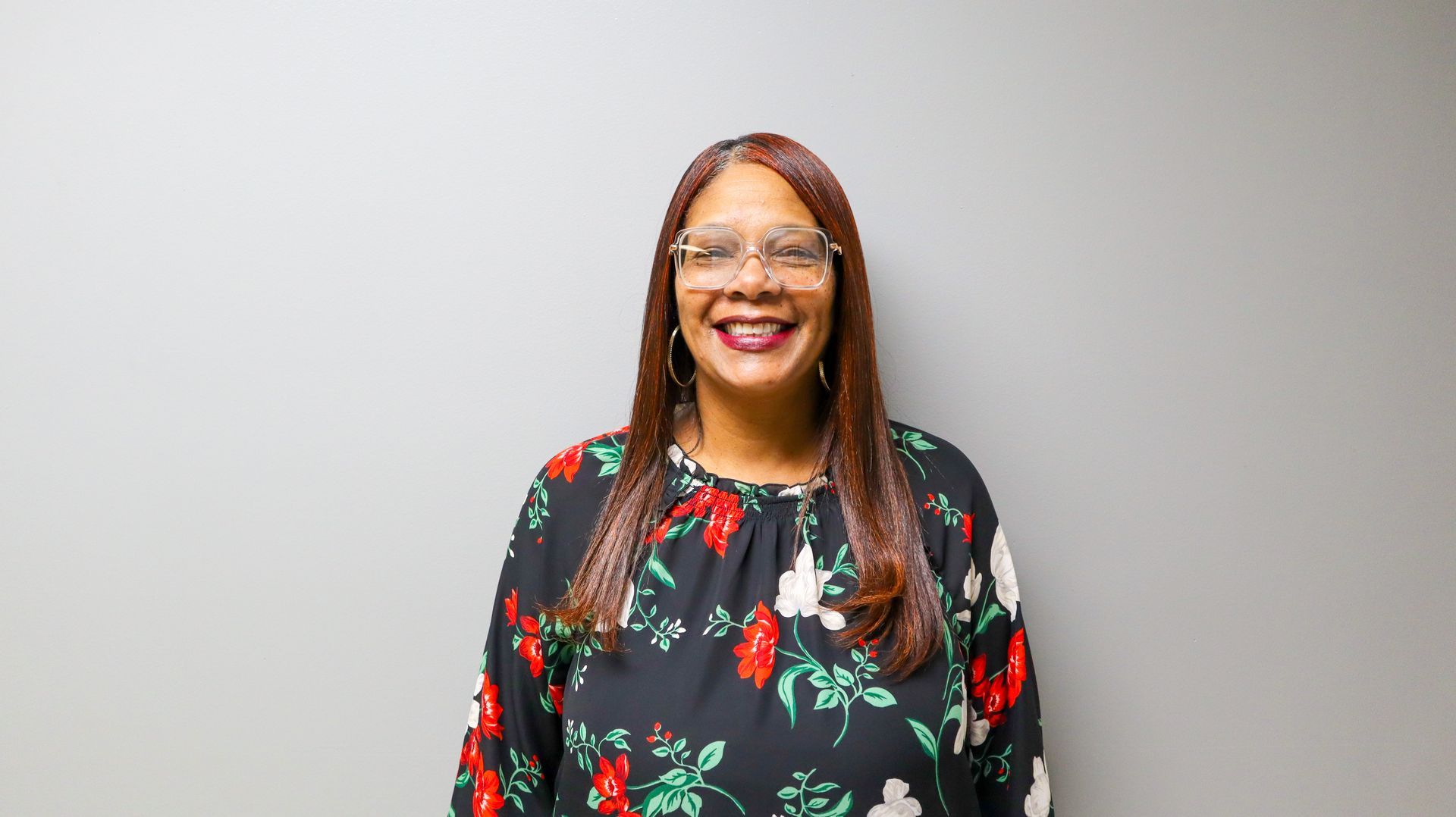 Smiling person with long hair and glasses wearing a black top with red and white floral patterns against a gray background.