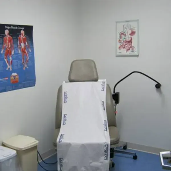 A medical examination room featuring a beige exam chair, a muscle anatomy chart, a wall print, and a trash bin.