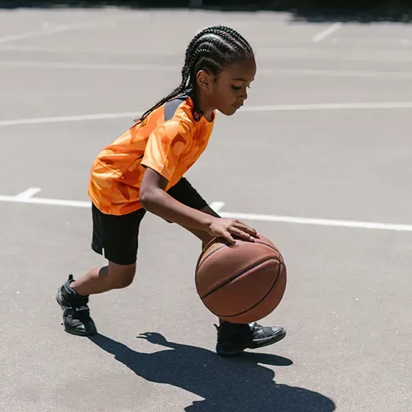 A person with braided hair wears an orange shirt and black shorts while dribbling a basketball on an outdoor court.