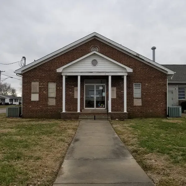 A red brick building with a gabled roof, a front portico with white columns, and a concrete walkway leading to the doors.