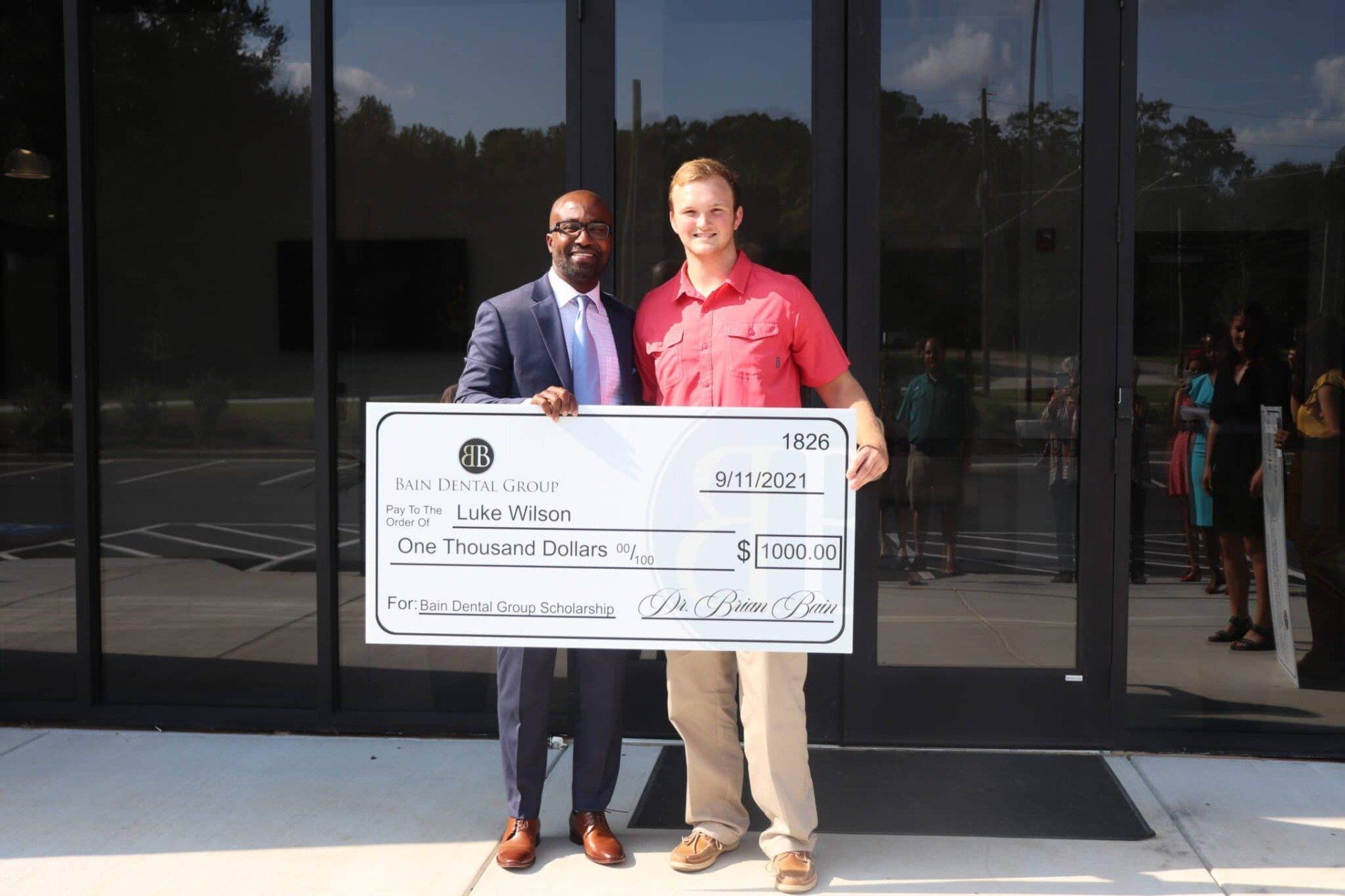 Two men are holding a large check in front of a building.