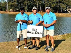 Three men holding golf clubs and a sign that says bain dental group