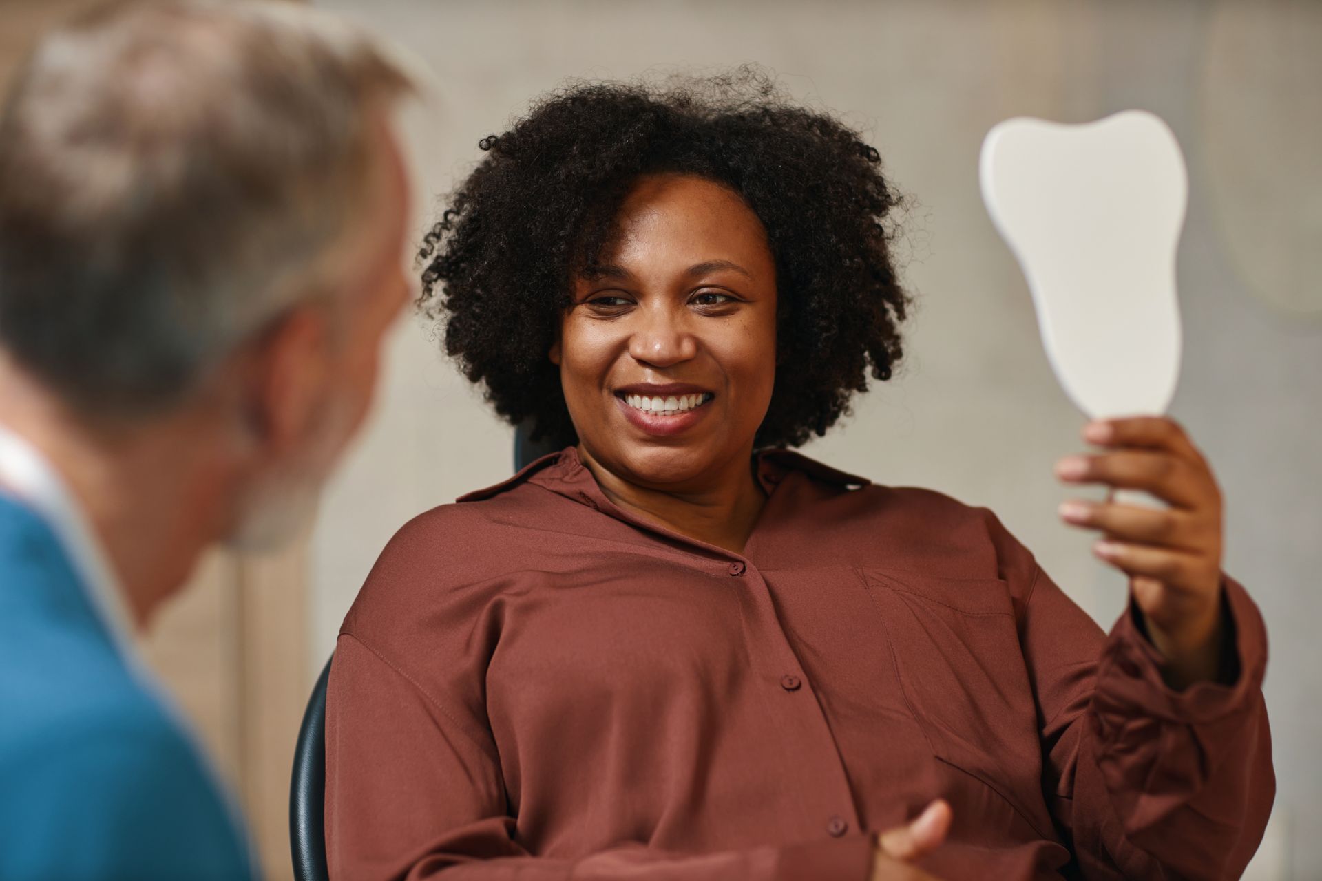A person holds a tooth-shaped shade guide while smiling, sitting across from a practitioner in a clinical setting.