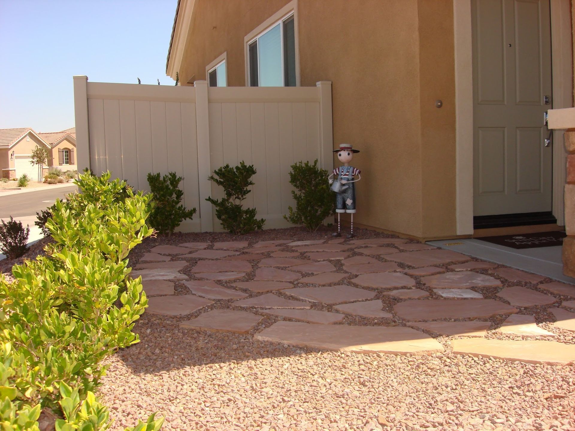 Beige house front with flagstone path, tan fence, green shrubs, and a decorative figure.